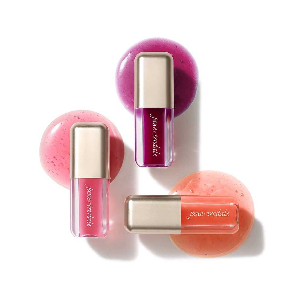 Drei jane iredale Just Kissed Lip Oils in Pink-, Beerenton- und Korallnuancen auf weißem Hintergrund, jeweils vor einem passenden glänzenden Farbswatch.