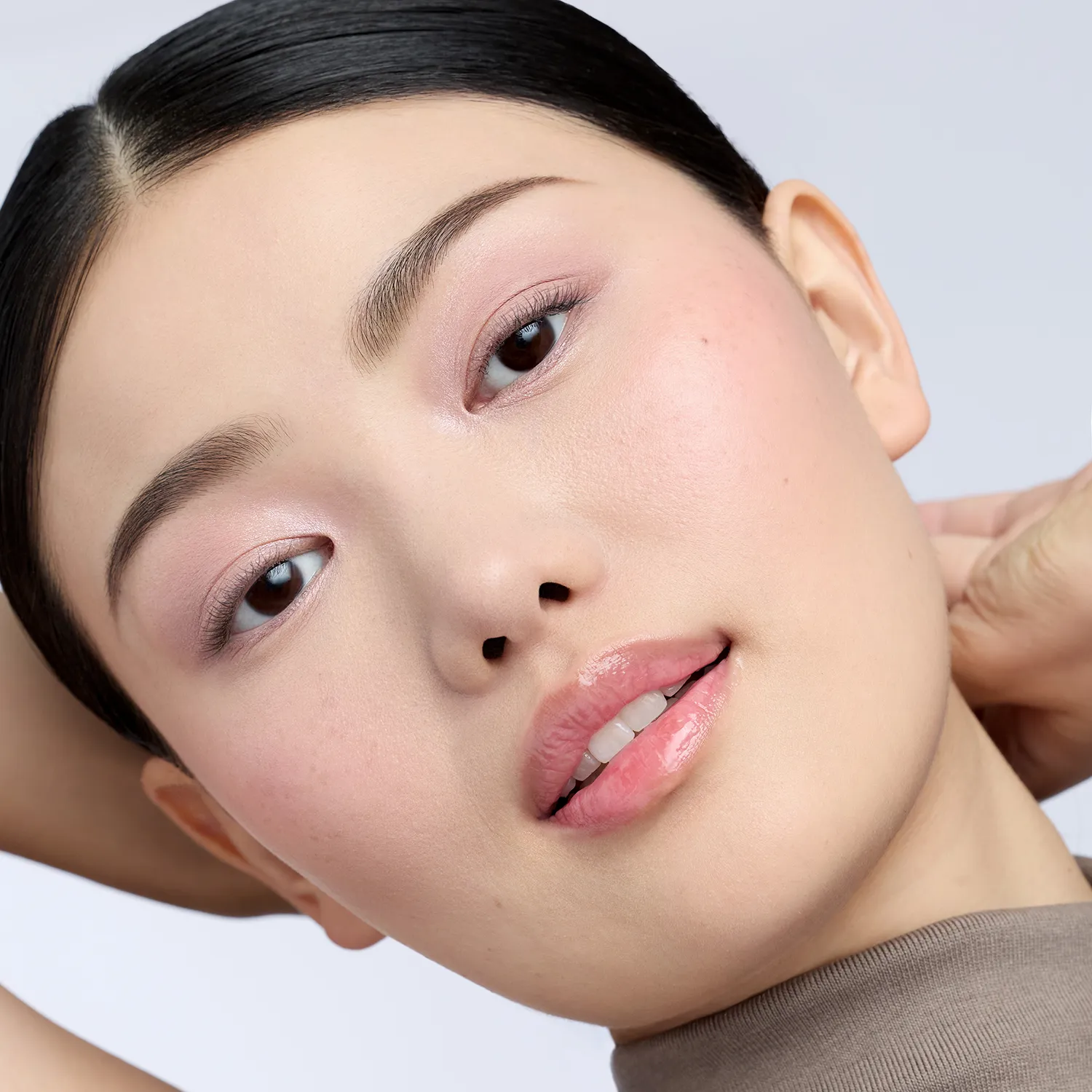 Nahaufnahme eines Gesichts mit dezentem roséfarbenem Augen-Make-up, glänzenden Lippen und sanft hervorgehobenen Wangen. Die Person hält beide Hände hinter dem Kopf.