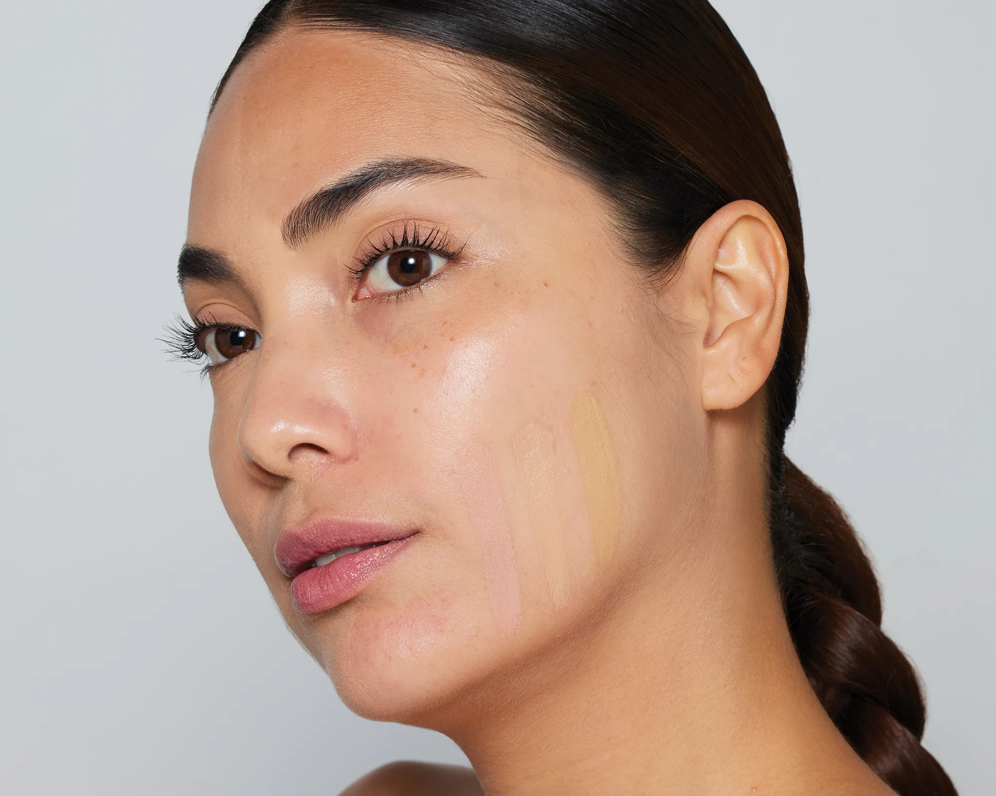Porträt einer Person mit zurückgebundenem dunklem Haar und dezentem Make-up. Auf der rechten Wange sind mehrere vertikale Foundation-Farbtests in unterschiedlichen Nuancen aufgetragen.