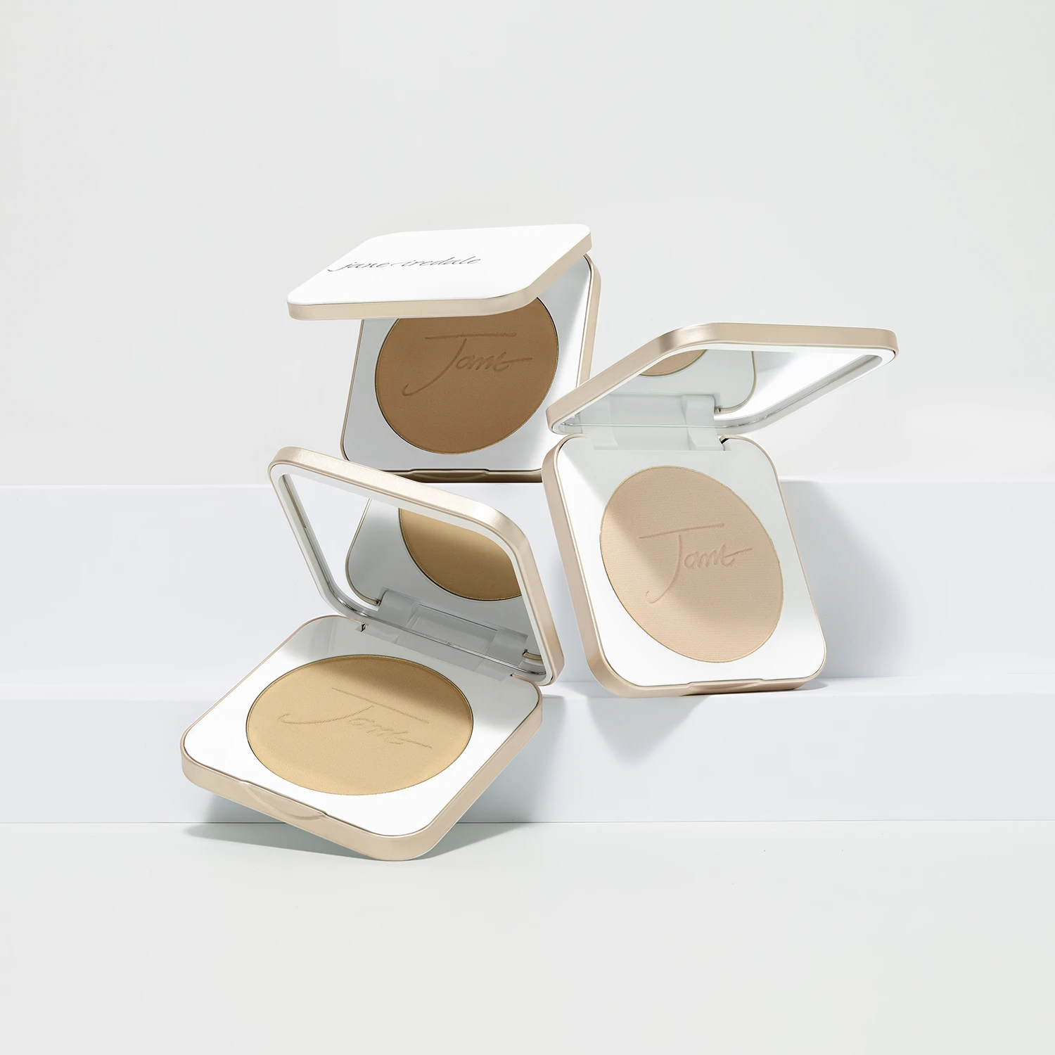 Drei geöffnete jane iredale PurePressed Base Mineral Foundations in unterschiedlichen Pudernuancen von hell bis dunkel, angeordnet in weiß-goldenen Kompaktdosen mit integriertem Spiegel auf hellem Hintergrund.
